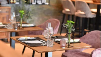 Ein Restaurant mit Holztischen und Stühlen, mit einer Vase mit Blumen, Weingläsern und Besteck auf dem Tisch. Die Stühle sind rosa und die Wände sind aus Stein.
