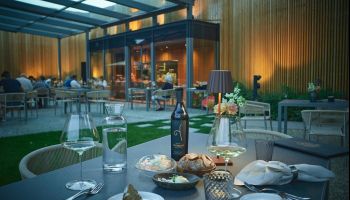 Ein Restaurant im Freien mit Glasdach, Holzwänden und warmer Beleuchtung. Ein Tisch ist mit Weingläsern, Tellern und einer Flasche Wein gedeckt. Im Hintergrund sitzen Menschen an anderen Tischen.