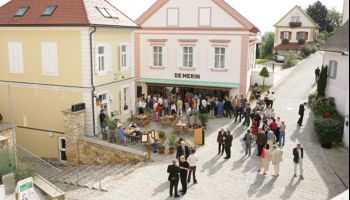 Eine Vogelperspektive auf einen Stadtplatz mit einer Menschenmenge vor einem Restaurant namens 'De Merin'. Das Gebäude hat eine rosa Fassade und einen grünen Vorbau. Menschen sitzen an Tischen und stehen, einige gehen auf der Kopfsteinpflasterstraße.