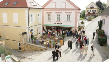 Eine Vogelperspektive auf einen Stadtplatz mit einer Menschenmenge vor einem Restaurant namens 'De Merin'. Das Gebäude hat eine rosa Fassade und einen grünen Vorbau. Menschen sitzen an Tischen und stehen, einige gehen auf der Kopfsteinpflasterstraße.