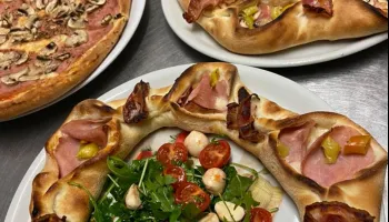 Drei Teller mit Pizza auf einem Tisch. Ein großer runder Pizza mit Schinken, Käse und Gemüse. Ein weiterer Teller mit einer kleineren Pizza mit Schinken und Käse. Ein dritter Teller mit einer Pizza mit Champignons und Käse.