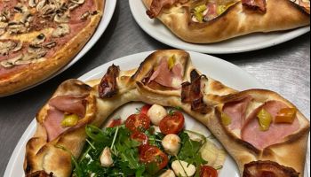 Drei Teller mit Pizza auf einem Tisch. Ein großer runder Pizza mit Schinken, Käse und Gemüse. Ein weiterer Teller mit einer kleineren Pizza mit Schinken und Käse. Ein dritter Teller mit einer Pizza mit Champignons und Käse.