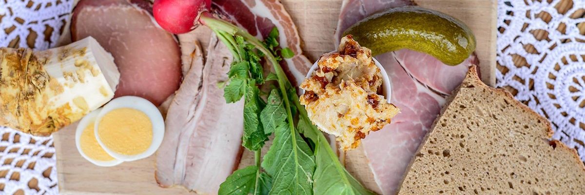 Ein Holzbrett zeigt eine Auswahl an Lebensmitteln, darunter Fleischscheiben, ein gekochtes Ei, eine Radieschen, ein Gewürzgurke und eine Schüssel mit Käseaufstrich mit einem Löffel.