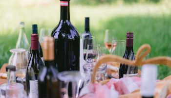 Ein Picknick-Setup mit verschiedenen Weinflaschen, Weingläsern und einem Korb auf einem Grasfeld. Die Flaschen sind in einer Reihe angeordnet, und einige sind geöffnet.