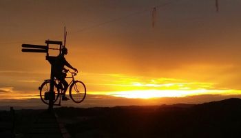 Eine Silhouette einer Person, die bei Sonnenuntergang ein Fahrrad mit einem Holzschild auf einem Pfosten fährt.