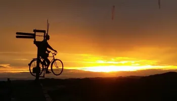 Eine Silhouette einer Person, die bei Sonnenuntergang ein Fahrrad mit einem Holzschild auf einem Pfosten fährt.