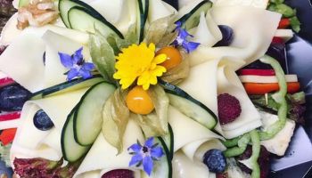 Ein Nahaufnahme eines Käseplatten mit Gurkenscheiben, Blaubeeren, Himbeeren und einer gelben Blume in der Mitte. Die Platte ist in einem kreisförmigen Muster auf einer schwarzen Oberfläche angeordnet.