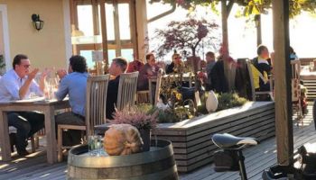 Eine Gruppe von Menschen sitzt an Tischen in einem Outdoor-Restaurant, mit einem Fahrrad und einem Fass in der Nähe. Die Umgebung ist sonnig mit Pflanzen und Bäumen im Hintergrund.