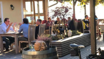 Eine Gruppe von Menschen sitzt an Tischen in einem Outdoor-Restaurant, mit einem Fahrrad und einem Fass in der Nähe. Die Umgebung ist sonnig mit Pflanzen und Bäumen im Hintergrund.