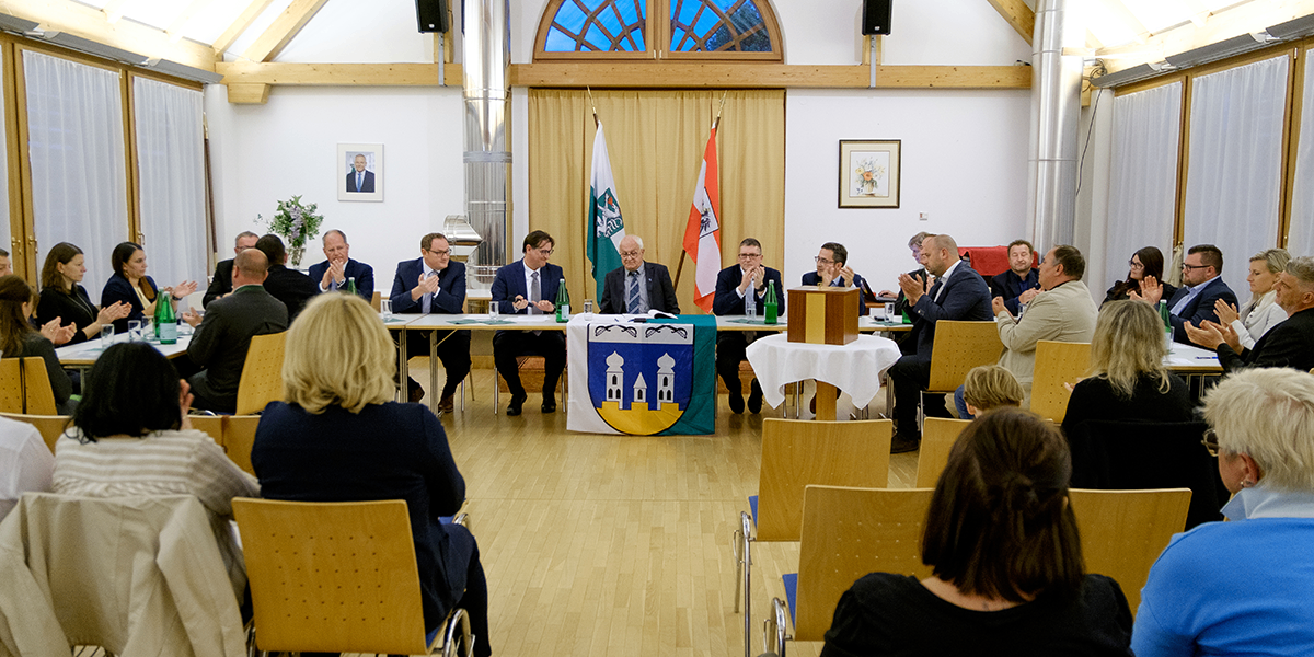 Eine Gruppe von Menschen in formeller Kleidung sitzt an einem Tisch in einem Besprechungsraum, mit Flaggen und einem Wappen im Hintergrund. Sie scheinen in eine Diskussion vertieft zu sein.