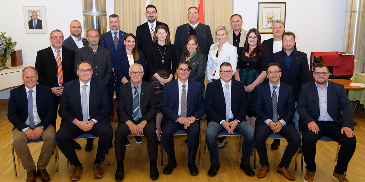 Eine Gruppe formell gekleideter Personen, sowohl Männer als auch Frauen, posiert für ein Foto in einem Raum mit Holzboden. Sie sitzen und stehen vor einer Kulisse mit einer Flagge und einem Bilderrahmen.