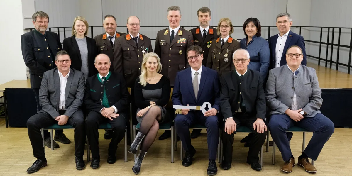 Eine Gruppe von Menschen in formeller Kleidung, einschließlich Militäruniformen, posiert für ein Foto. Ein Mann vorne hält einen Schlüssel.