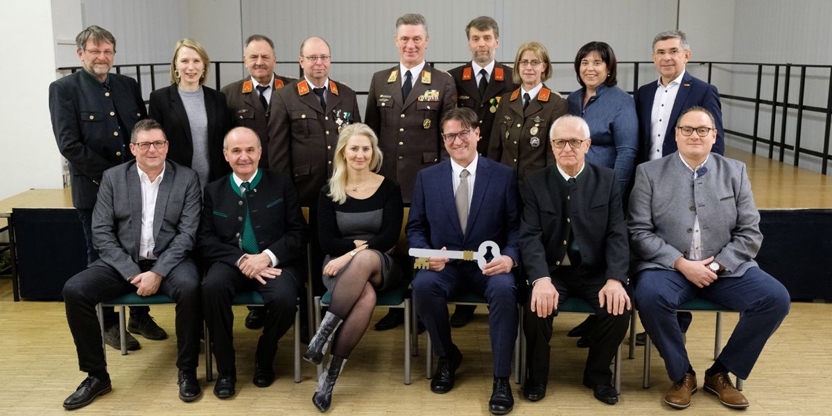 Eine Gruppe von Menschen in formeller Kleidung, einschließlich Militäruniformen, posiert für ein Foto. Ein Mann vorne hält einen Schlüssel.
