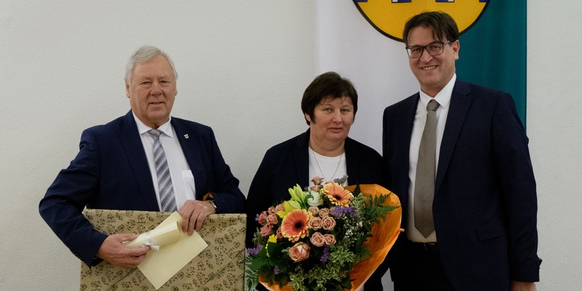 Drei Personen stehen zusammen, eine hält einen Blumenstrauß, eine andere ein eingewickeltes Geschenk und die dritte lächelt. Sie sind formell gekleidet.