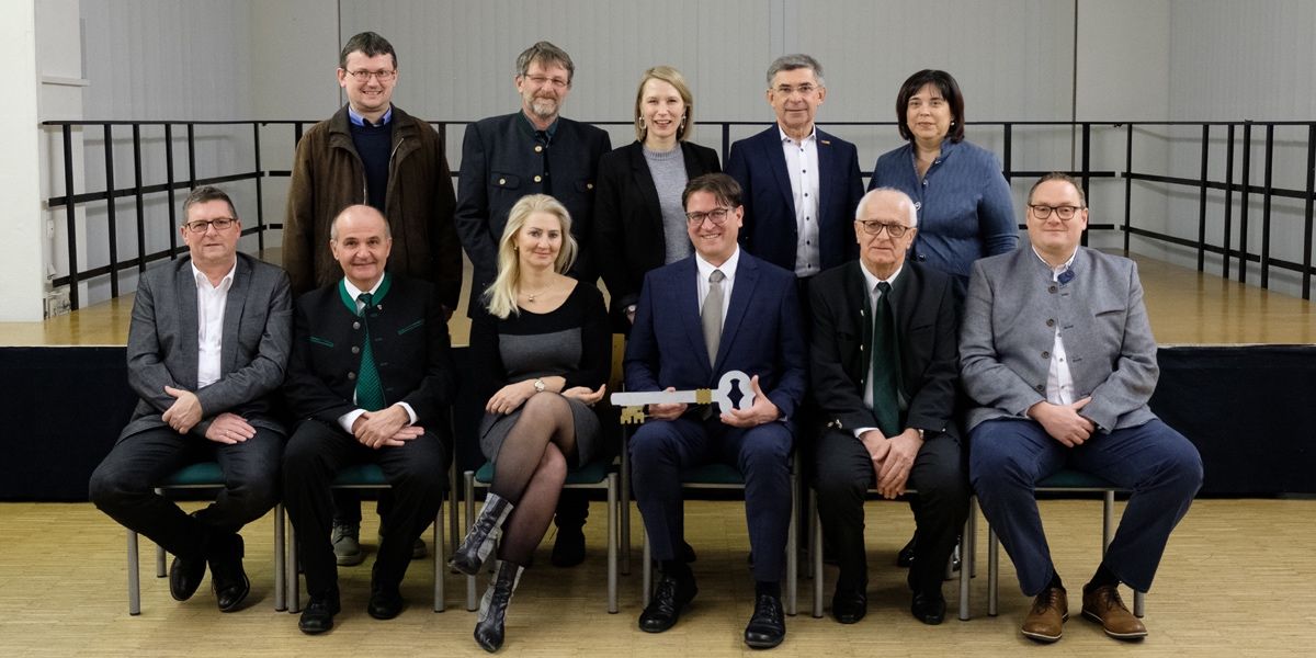 Eine Gruppe formell gekleideter Personen, einschließlich Männer und Frauen, sitzen auf Stühlen und lächeln für ein Foto. Ein Mann hält einen großen Schlüssel.