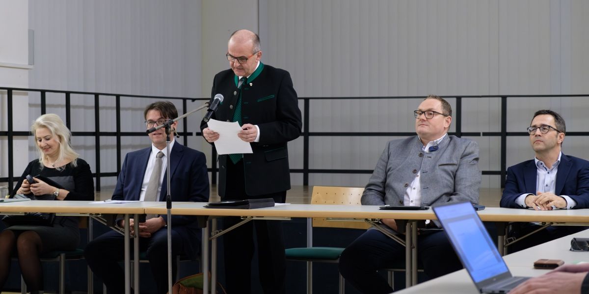 Drei Männer in Anzügen sitzen an einem Tisch, einer steht und hält ein Stück Papier in der Nähe eines Mikrofons. Der stehende Mann liest aus dem Papier.