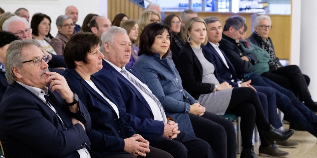 Eine Gruppe von Menschen in formeller Kleidung sitzt in einer Reihe, möglicherweise bei einer Konferenz oder Besprechung. Die Frau in der Mitte schaut nach vorne, während andere in verschiedene Richtungen schauen.