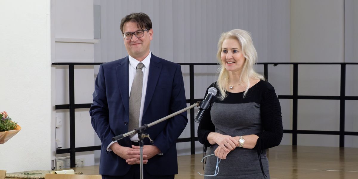Ein Mann und eine Frau stehen lächelnd nebeneinander vor einem Mikrofon auf einem Ständer. Der Mann trägt einen Anzug und eine Krawatte, die Frau ein schwarzes und graues Kleid. Hinter ihnen befindet sich ein Metallgeländer.