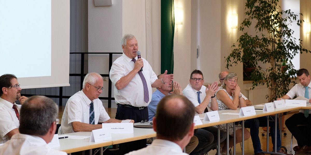 Eine Gruppe von Menschen sitzt an einem Tisch und hört einem älteren Mann zu, der in ein Mikrofon spricht. Der Mann ist in ein weißes Hemd und eine lila Krawatte gekleidet.