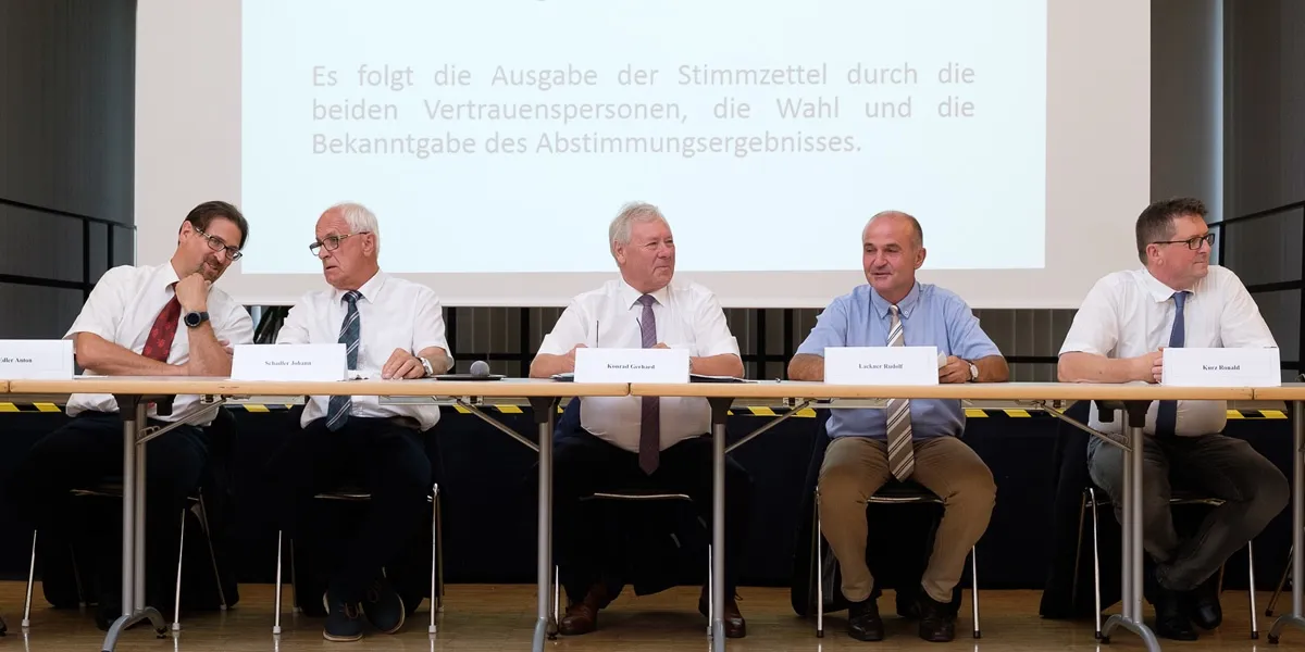 Vier Männer sitzen in einem Konferenzraum an einem Tisch, vor ihnen ein Beamer, der deutschen Text anzeigt. Sie sind in formeller Kleidung gekleidet, mit Namensschildern vor ihnen.