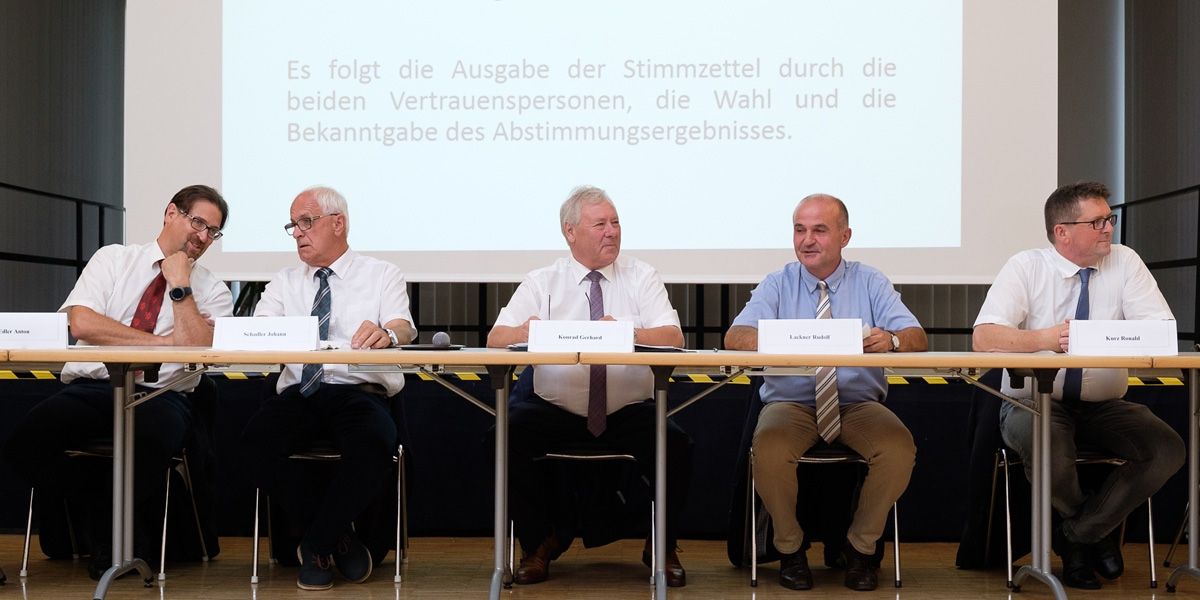 Vier Männer sitzen in einem Konferenzraum an einem Tisch, vor ihnen ein Beamer, der deutschen Text anzeigt. Sie sind in formeller Kleidung gekleidet, mit Namensschildern vor ihnen.
