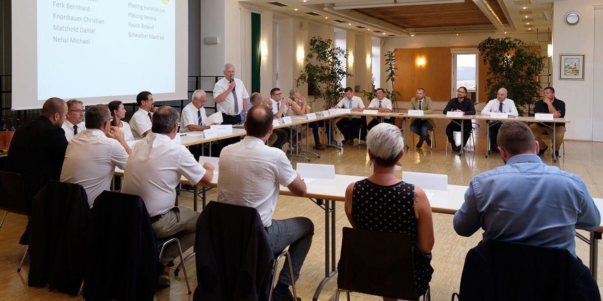 Eine Gruppe von Menschen in formeller Kleidung sitzt um einen Tisch in einem Konferenzraum herum, wobei ein Mann an einem Podium steht und in ein Mikrofon spricht. Eine Projektionsleinwand ist im Hintergrund sichtbar.