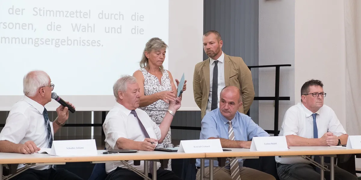 Eine Gruppe von Menschen sitzt an einem Tisch, möglicherweise in einer Besprechung. Eine Frau steht hinter ihnen und hält ein Stück Papier. Der Mann auf der linken Seite hält einen Stift. Der Mann in der Mitte trägt eine Krawatte. Der Mann auf der rechten Seite trägt eine Uhr.