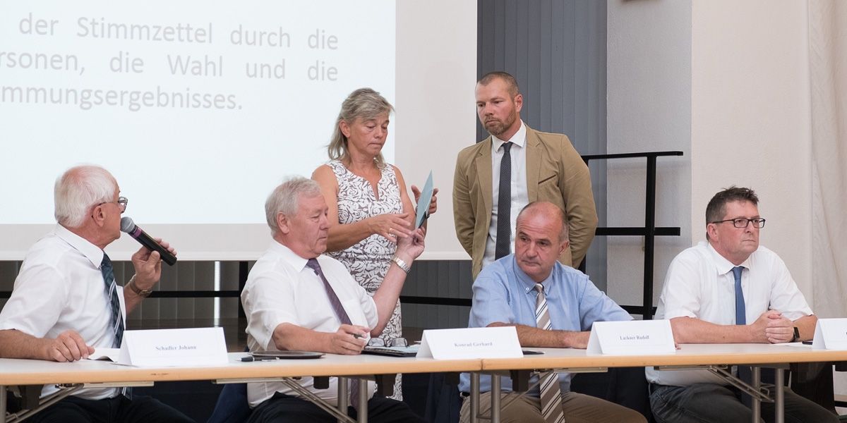 Eine Gruppe von Menschen sitzt an einem Tisch, möglicherweise in einer Besprechung. Eine Frau steht hinter ihnen und hält ein Stück Papier. Der Mann auf der linken Seite hält einen Stift. Der Mann in der Mitte trägt eine Krawatte. Der Mann auf der rechten Seite trägt eine Uhr.