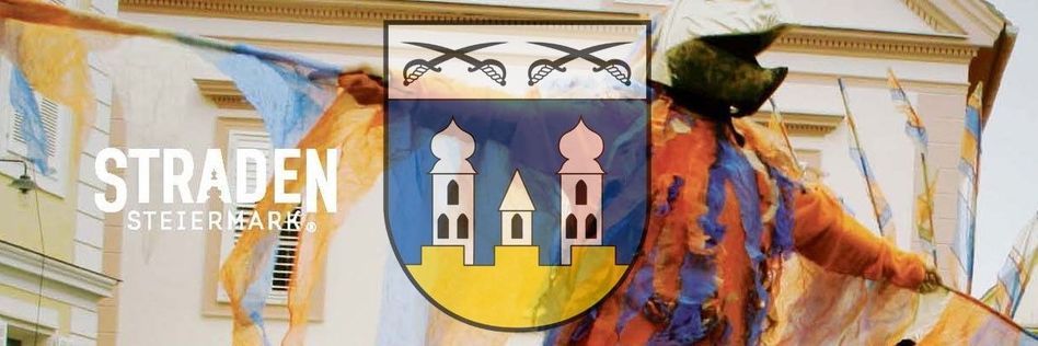 Das Bild zeigt ein farbenfrohes Wappen mit zwei Schwertern und drei Türmen, das auf einem Banner abgebildet ist. Eine Person in einem Kostüm steht neben dem Wappen und hält es. Der Hintergrund umfasst ein Gebäude mit einem Fenster und einem dekorativen Muster.