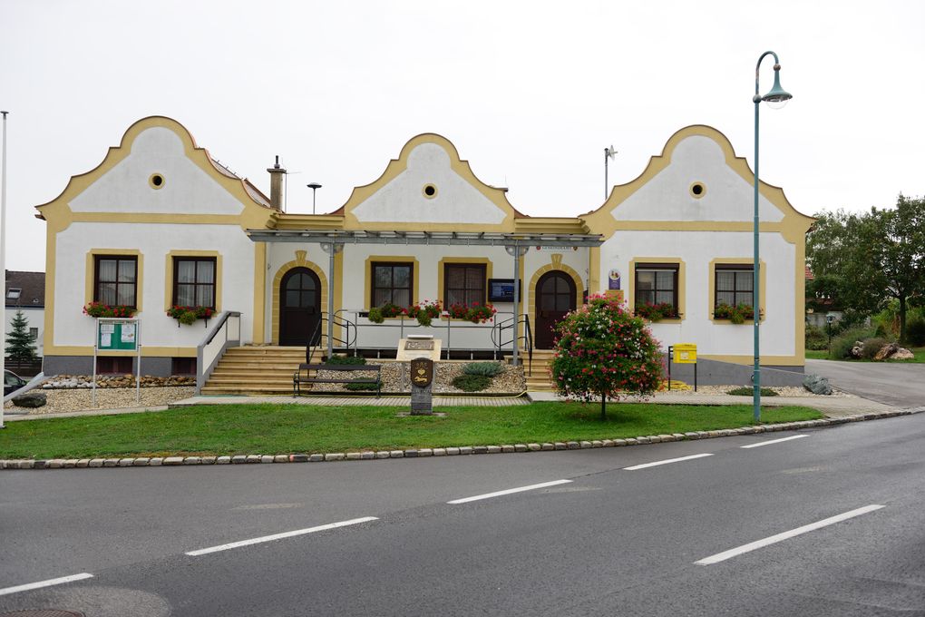  Abschnitt Bild: Ein dreistöckiges Gebäude mit einer gelb-weißen Fassade hat drei Türen und mehrere Fenster. Eine Bank und ein Blumenbeet stehen davor. Eine Straße mit weißen Markierungen befindet sich vor dem Gebäude.