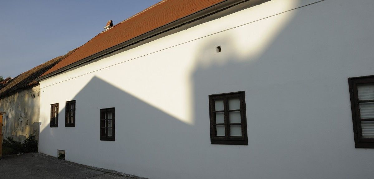 Ein weißes Gebäude mit einem braunen Dach und zwei braun gerahmten Fenstern. Ein Schatten des Gebäudes fällt auf die Wand.
