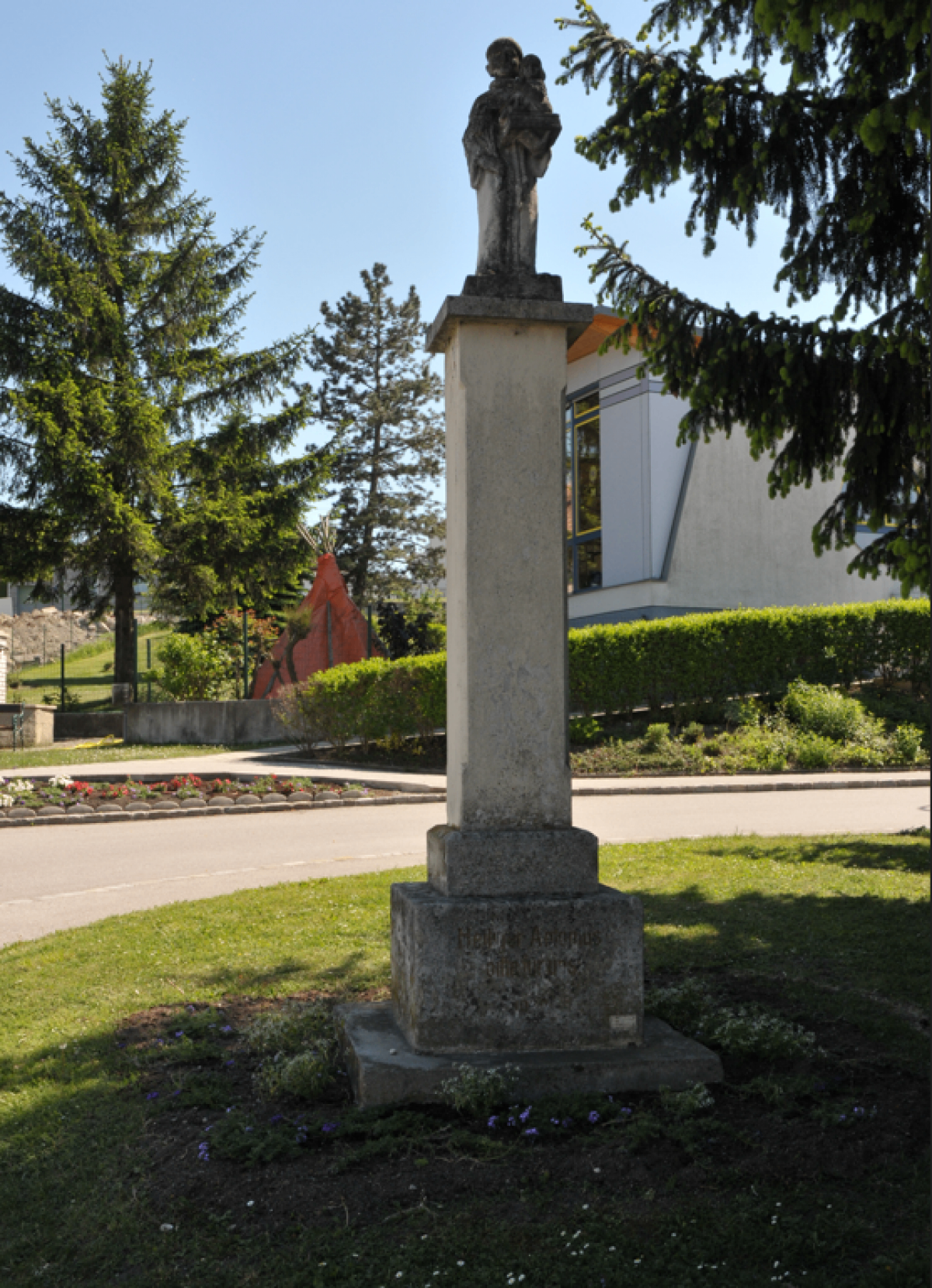 Ein hoher Steinmonument steht in einem Park mit üppigen grünen Bäumen und Büschen. Dahinter befindet sich ein Gebäude mit Glasfenstern und einem roten Zelt. Das Gebiet ist gut gepflegt.