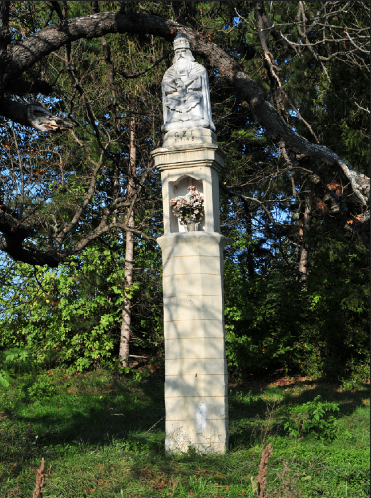 Ein hoher Steinmonument mit einer sitzenden Statue auf der Spitze, datiert 1765, steht in einem Wald. Es hat eine kleine Blumenarrangement an seiner Basis.