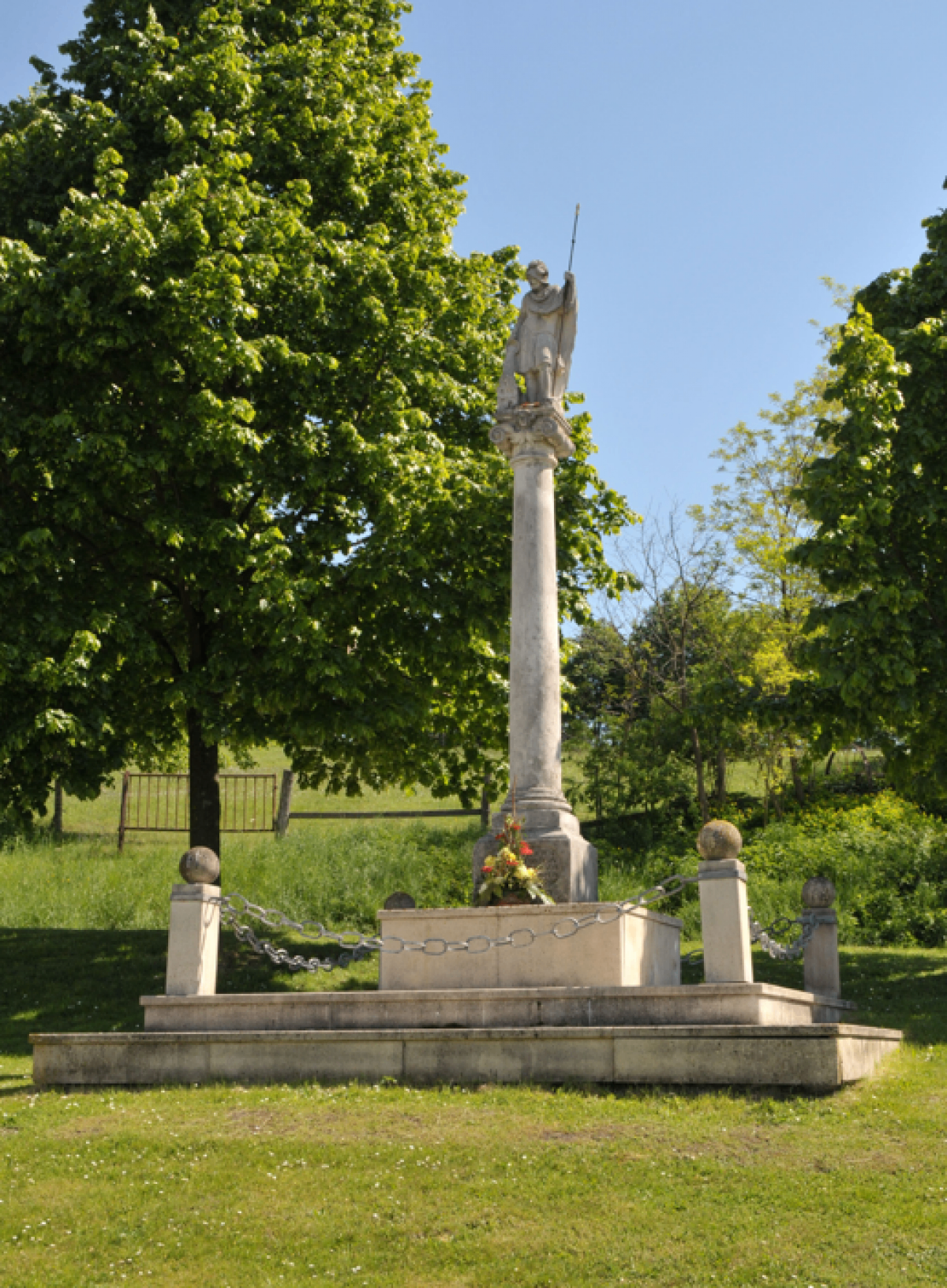 Eine Statue eines Soldaten steht auf einem Steinpfeiler in einem Park, umgeben von einem Kettenzaun. Bäume und ein Zaun sind im Hintergrund.