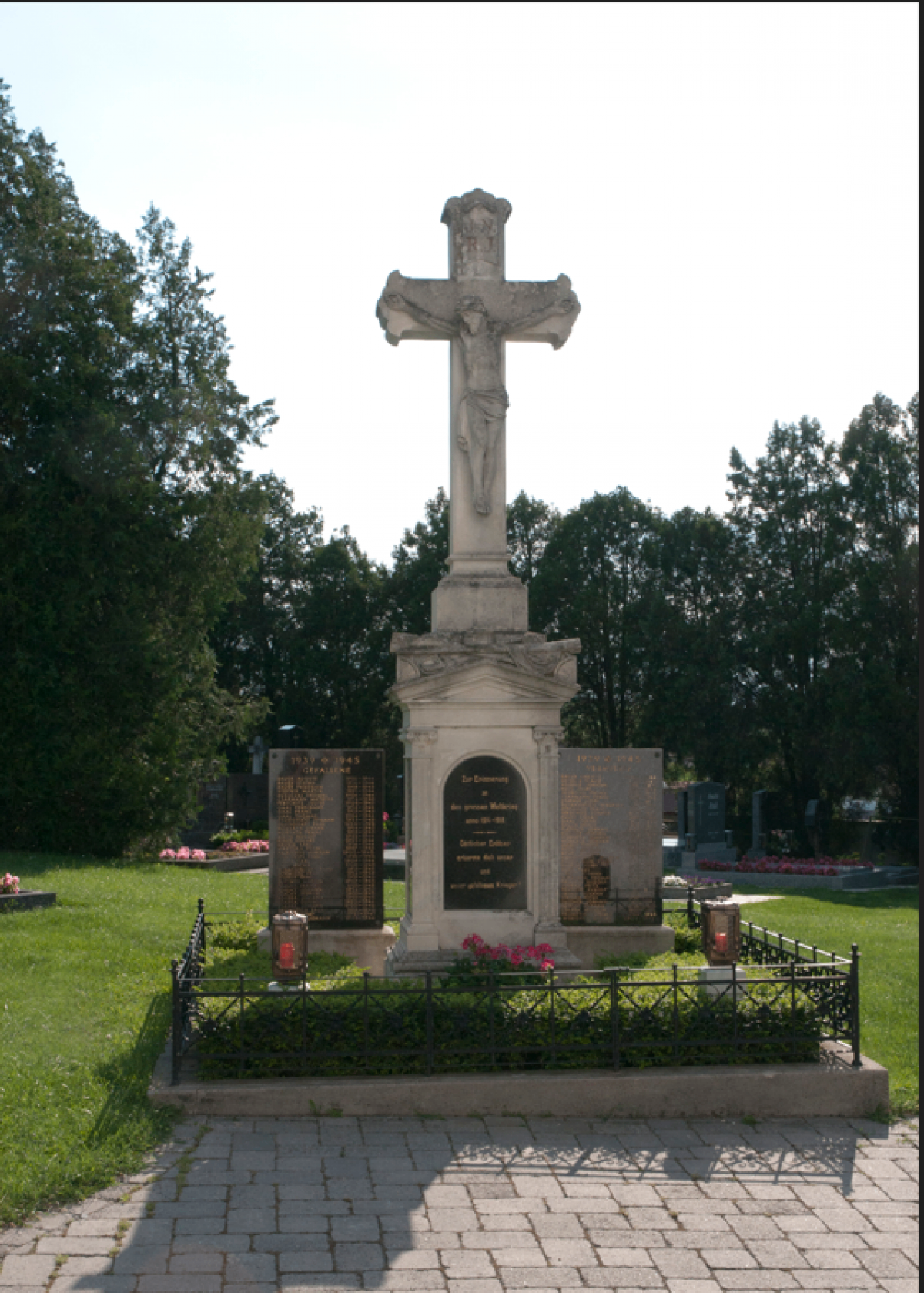 Ein Gedenkkreuz steht auf einem Friedhof. Es zeigt einen Kruzifix mit Jesus Christus und zwei Tafeln mit Inschriften. Es ist von Grünpflanzen und Blumen umgeben.
