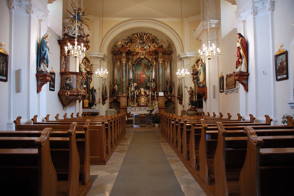 Ein leerer Kircheninnenraum mit hölzernen Kirchenbänken, Altären und Skulpturen. Ein zentraler Gang führt zu einem großen Altarbild und Statuen an den Wänden.