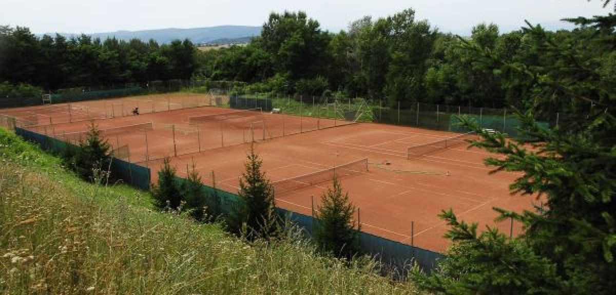 Im Freien gelegene Tennisplätze umgeben von Bäumen und Gras, mit einer Person in der Nähe der Plätze sichtbar.