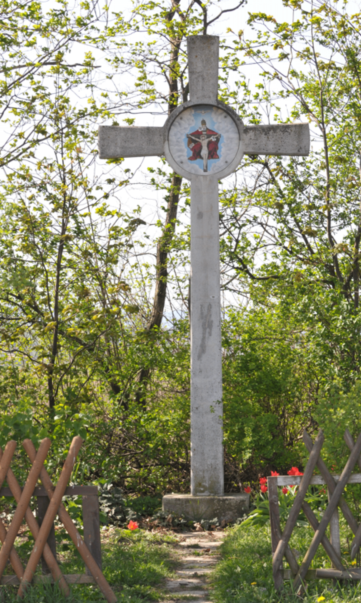Ein hoher Betonkreuz steht in einem Wald mit einem roten Kruzifix-Bild. Bäume umgeben das Kreuz.