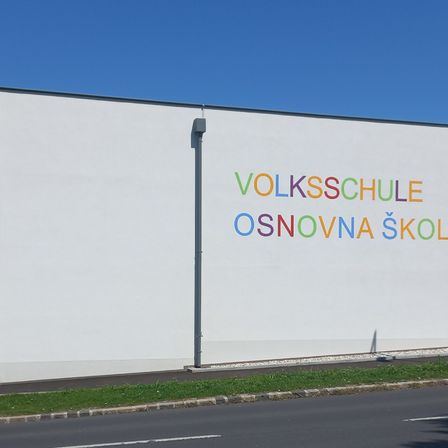 Eine weiße Wand mit bunter Schrift darauf zeigt 'Volksschule Osnovna Skol'. Der Himmel ist blau und klar.