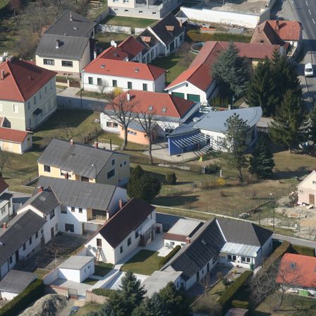 Luftaufnahme eines Wohngebietes mit mehreren Häusern und Gebäuden, unterschiedlichen Dachfarben und -stilen, umgeben von Bäumen und Grünflächen. Eine Straße und ein Fahrzeug sind in der Ferne zu sehen.