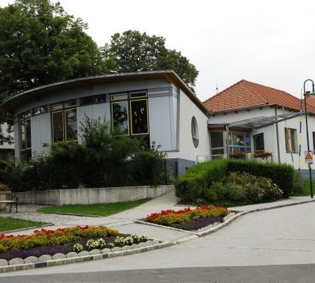 Ein weißes Gebäude mit roten Dächern und Fenstern. Das Gebäude ist von Blumen und Pflanzen umgeben. Es gibt eine Bank und eine Laterne an der Seite.