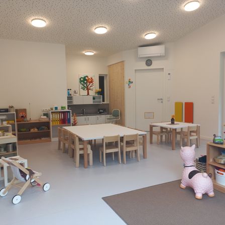 Ein gut beleuchteter Raum mit weißen Wänden und Decken. Er hat einen weißen Tisch und Stühle, eine weiße Tür und einen Teppich. Es gibt Regale mit Spielzeug und Büchern. An der Wand befinden sich eine Uhr und eine Klimaanlage.