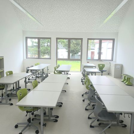 Ein leerer Klassenraum mit weißen Wänden und Tischen. Grüne Stühle sind in Reihen aufgestellt. Drei Fenster bieten Blicke auf Bäume und ein saftiges Feld.