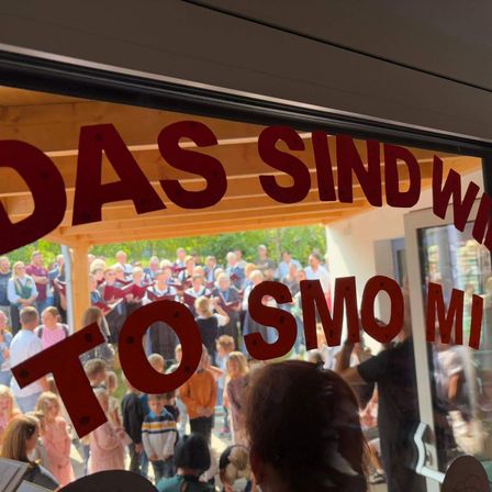 Eine Menschenmenge versammelt sich vor einem Gebäude, einige halten rote Hefte in der Hand. Ein Schild auf der Scheibe lautet 'TO DAS SIND SMO MI'.