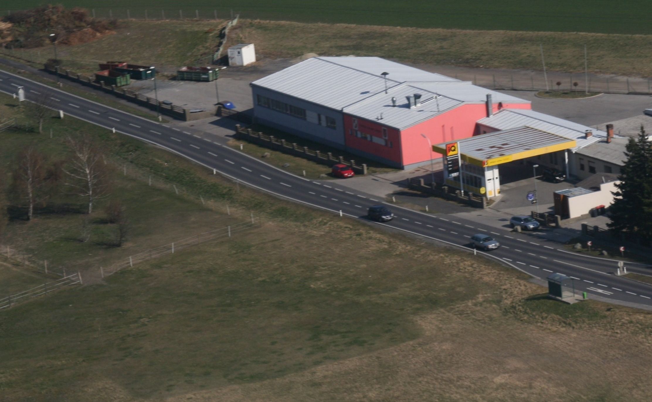 Eine Luftaufnahme eines ländlichen Gebiets mit einer Straße, einem roten Auto, das in der Nähe eines Gebäudes geparkt ist, einer Tankstelle und einem Feld.