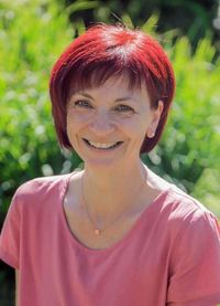Eine Frau mit kurzen roten Haaren lächelt und steht in einem Garten. Sie trägt ein rosa Oberteil und eine Halskette mit einem kleinen orangefarbenen Anhänger.