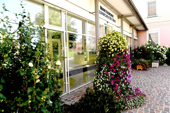 Ein Gebäude mit Glastüren und Fenstern hat eine große, farbenfrohe Blumenskulptur davor. Das Gebäudeschild lautet 'Marktgemeinden Eidkirchen bei Graz'.