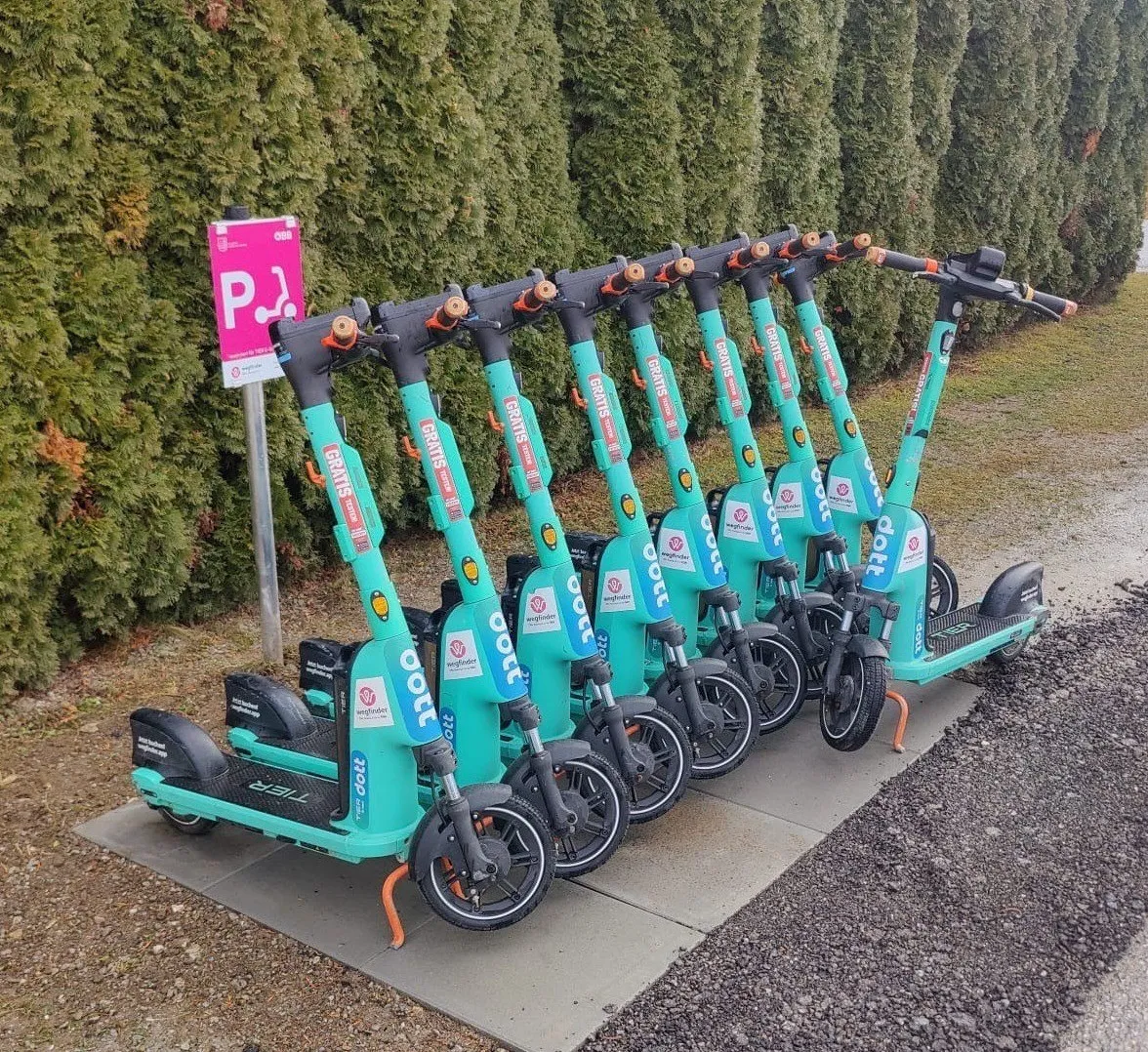 Mehrere türkisfarbene Elektroroller sind in einer Reihe auf einem Bürgersteig geparkt, mit einem Parkzeichen in der Nähe.