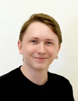 Eine Person mit kurzen braunen Haaren und blauen Augen, die ein schwarzes T-Shirt trägt, lächelt und scheint für ein Foto zu posieren.