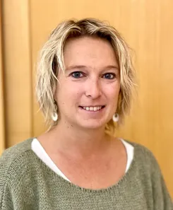 Eine Frau mit blonden Haaren lächelt und steht vor einer Holzwand. Sie trägt einen grünen Pullover und weiße Ohrringe.